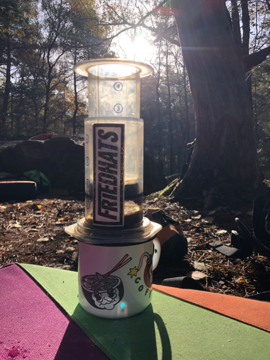 Image is outside in a park or forest with big trees surrounding. An AeroPress sits on top of a travel mug with a ramen bowl and bomb on it, cartoon style. There is a Friedhats coffee sticker on the side of the AeroPress.