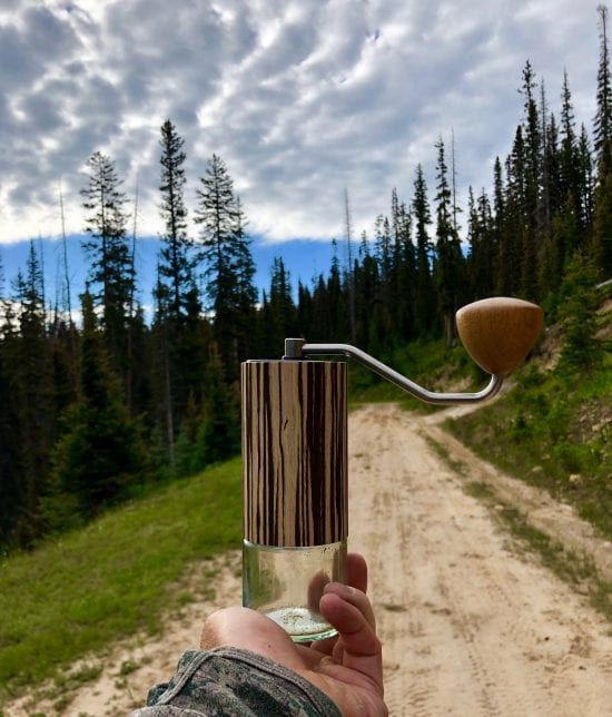 A hand holds up a wooden Comandante grinder. It has a large wooden knob on a long metal arm at the top, a wooden middle, and a clear glass bottom. In the background is a dirt trail leading into an evergreen forest.