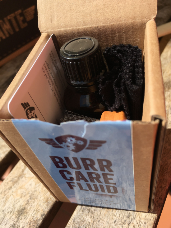 Inside the burr care fluid box. There is a glass bottle with a screw on lid, a flat wooden bristle brush, and a cleaning cloth.