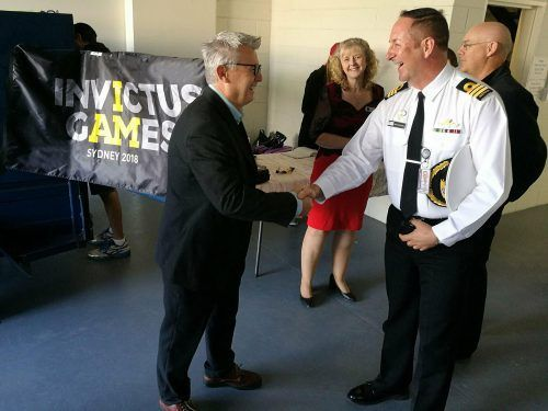 A gray-haired man in a suit, wearing glasses, shakes hands with a man in uniform. Onlookers smile near an Invictus Games banner.