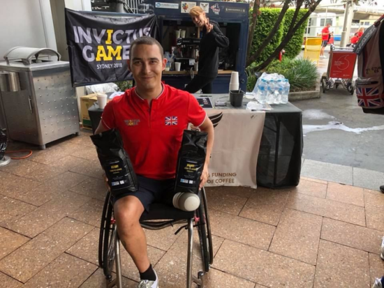 A veteran in a wheelchair, missing one leg,  holds up two bags of coffee near the 3 Elements coffee kiosk at the Invictus Games in Australia. Someone at the kiosk in the background gives a peace sign. 