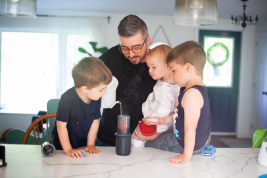 Justin and his three kids watch while he pours water from a white gooseneck kettle into an AeroPress. They are at the kitchen table with an overhead lamp. A couple of houseplants are visible, and a gray door and chandelier are in the background. 