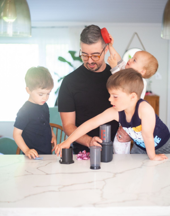 Justin brews coffee with his three small children in tow. They are in the kitchen, using an AeroPress at the table. The smallest child is trying to put a red toy on Justin's head.