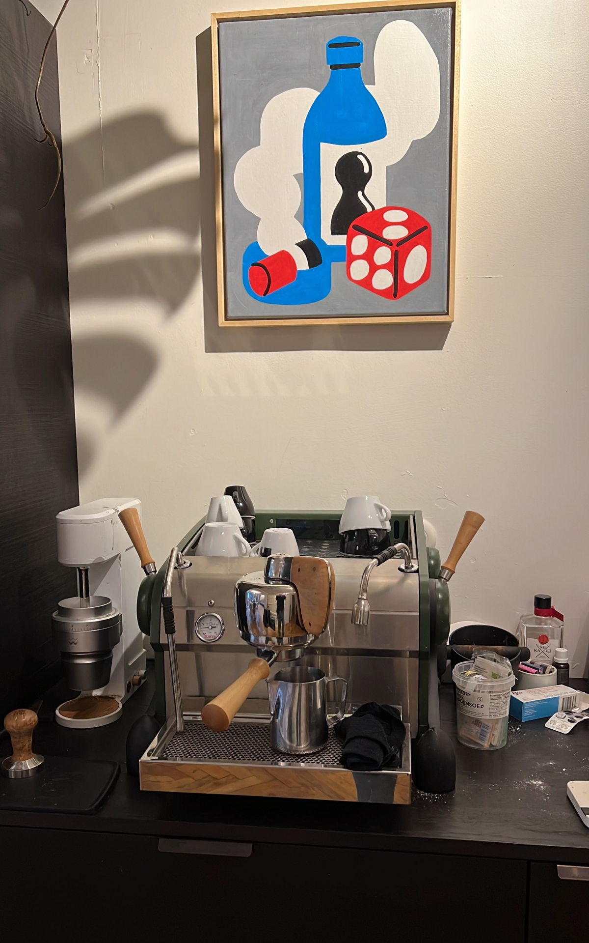 Lex's home espresso machine setup. He has a small espresso machine with wooden handles for the steam wands and a wooden paddle for the portafilter. Small cappuccino cups sit on top. To the left is a coffee brewer, and to the right is other coffee paraphernalia like sweeteners. On the white wall above is modern art depicting a blue bottle, a red die and what may be a lit cigarette.