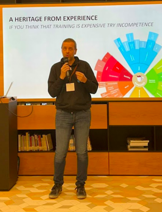 Edgardo speaks into a microphone at the Coffee Technicians Guild Summit in Italy in 2022. he wears a black pullover and jeans, and behind him is a large screen with a PowerPoint presentation slide that reads: A Heritage From Experience: If you think training is expensive, try incompetence.