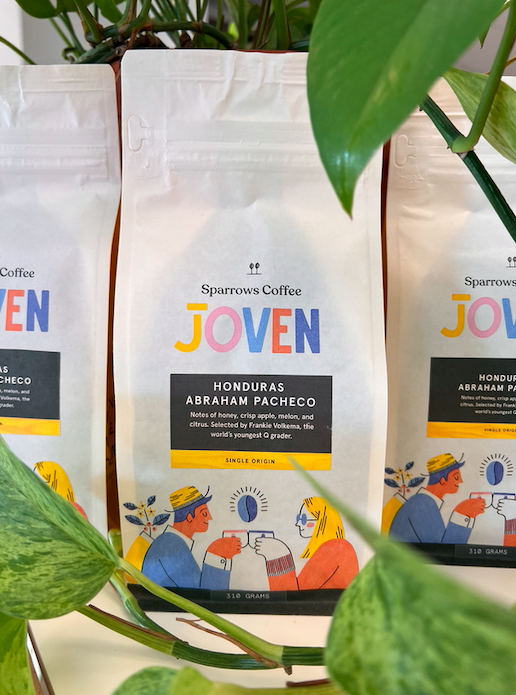 A bag of Joven coffee features an illustration of two people clinking coffee mugs together in a toast underneath a floating blue coffee bean. The coffee is Honduras Abraham Pacheco, with notes of honey, crisp apple, melon and citrus.