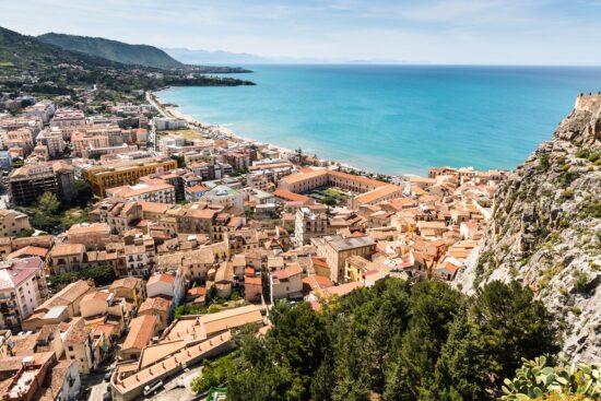 An overhead view of the Sicilian coast and a coastal town. On the right are rocky cliffs looming up, and hills on the left, with the red-roofed and tan buildings between in a large grotto. The beach is a bright white, and the ocean is a clean blue.