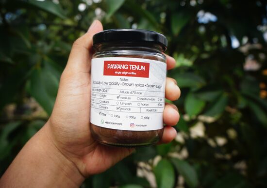 A clear glass jar of ground coffee being held in a woman's hand. The red label says Pawang Tenun single origin coffee blend and lists medium roast, with notes of brown sugar and spice and low acidity.