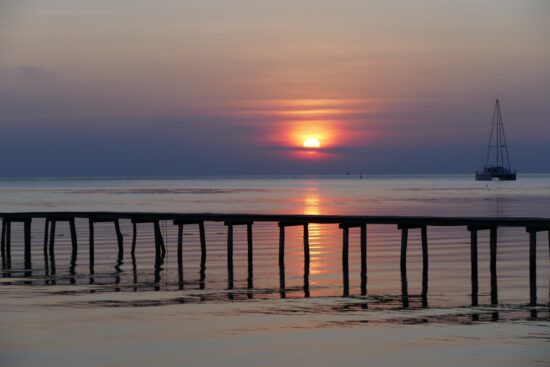 A simple wooden dock just out into water in Indonesia at sunset. The sky is an array of colors from pink and purple to orange. A small sailing boat is coming towards shore.