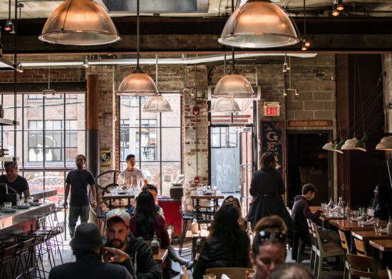 The interior of a bustling Toronto cafe. The space is large and industrial looking, with exposed brick, hanging lamps, and tall windows and a glass door. The tables are wood and metal, with plain chairs and a shiny espresso bar.  An assortment of people sit at the bar and at tables for two.