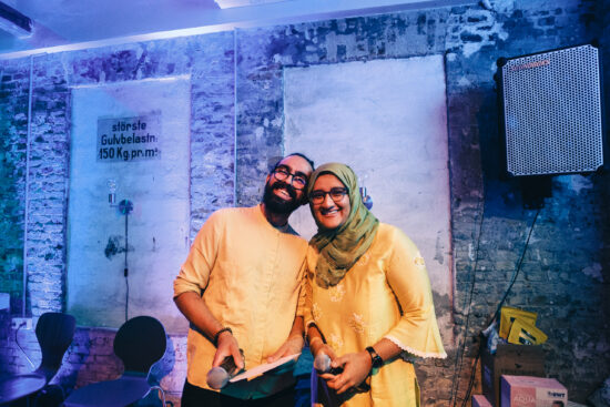 Two people in yellow shirts pose for the camera in Copenhagen. One wears glasses and a beard, the other glasses and a green head scarf. The background is a blue wall.
