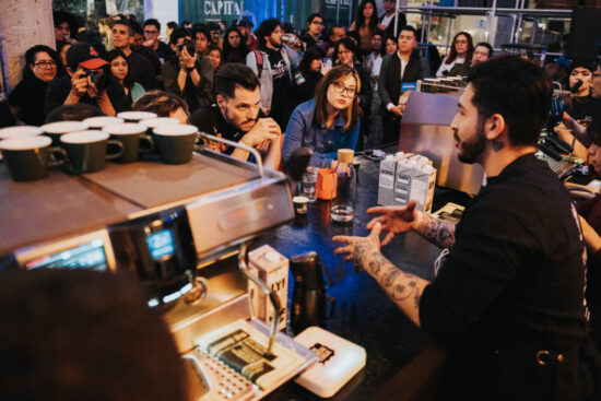 Jako tetovirani barista govori iza espresso bara u Mexico Cityju dok suci i gledatelji slušaju.