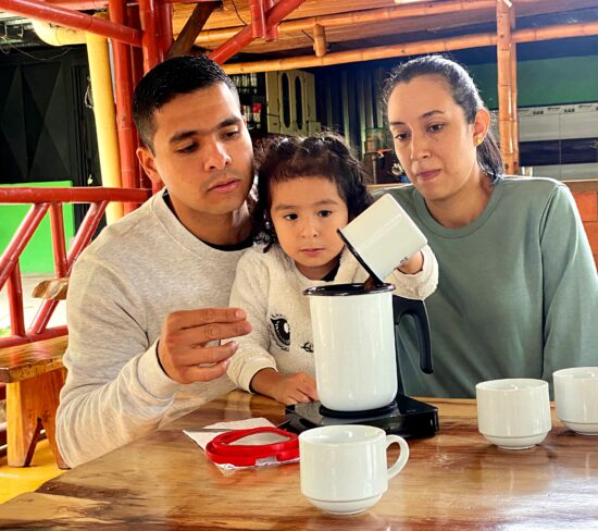 Diego with his wife and young daughter brewing coffee at home. His daughter is pouring coffee into a white pot with a handle while her parents look on.