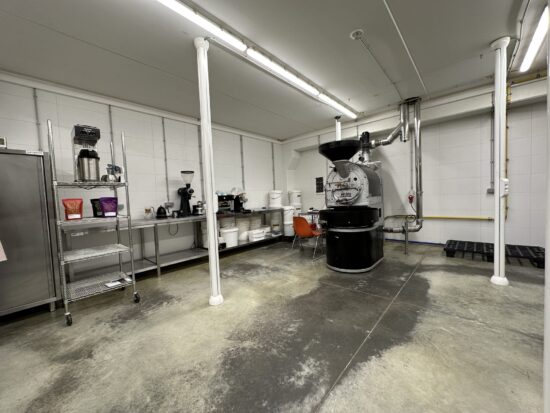 A basement type facility with a roaster and coffee equipment like grinders and tubs on metal shelves.