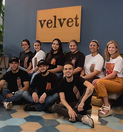 Six young women and three young men from Cafe Velvet pose for a group photo in front of a large sign that reads "velvet." The wall is purple and the floor is hexagon tiled.