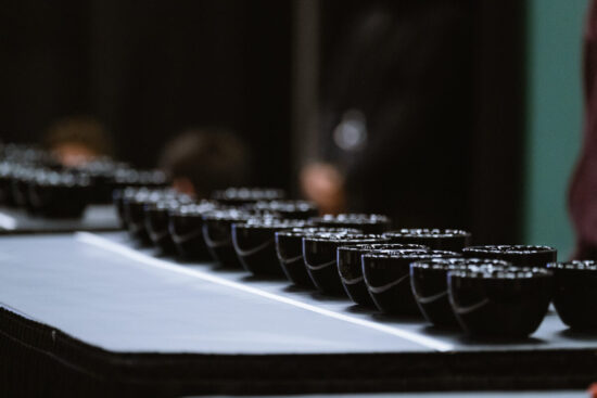 Rows of black cupping vessels are lined up on a long table for cup tasters to try.