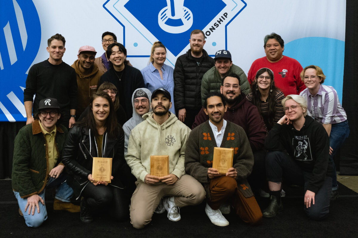 A group of 18 baristas pose for the winner photo, all of varying ages, gender identities and backgrounds. The front of the group kneels, and the top three winners hold up wooden plaques.