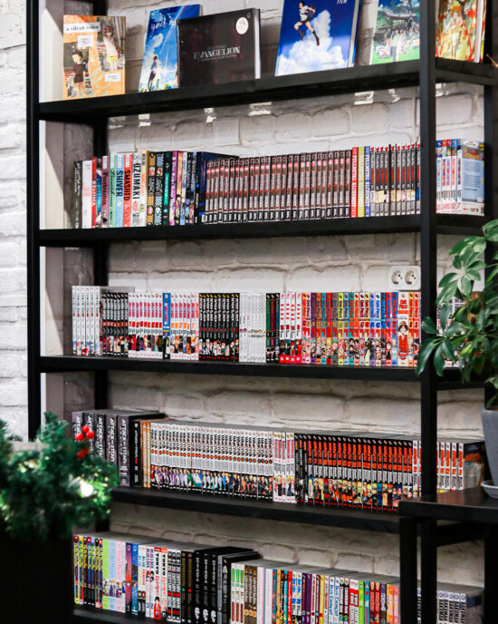 A large black bookshelf with an open back to the exposed white brick wall inside the shop. The shelves are packed with comics and manga, with some larger books facing out on top so you can see the covers. 