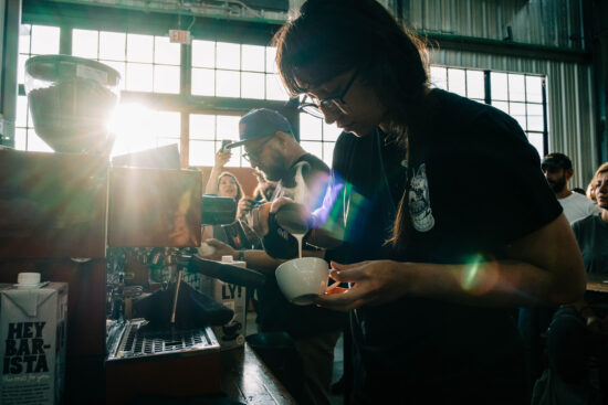 Two baristas at work on an espresso machine at the throwdown. One is a young woman in glasses and a braid, the other a man with glasses and a baseball cap.