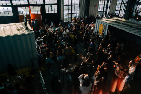 A crowd inside a warehouse area with metal storage crates and huge windows.