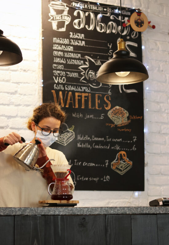 A barista performs a pour over into a Hario V60 drip. A menu board behind her displays drinks, waffle options, and cartoons drawn on it.