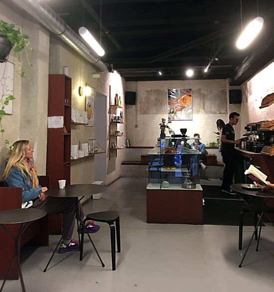 Customers sit inside Acid #1 while baristas make drinks. There is abstract art on the walls, small black tables with wooden benches on a concrete floor, and  glass pastry case. The ceiling is black with bright industrial lighting.
