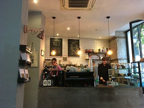 The baristas behinf the chic black and white bar at Hanso. There is a Strada machine and a glass pastry case, with whhite subway tile walls behind the bar and black shelving for coffee and merchandise.