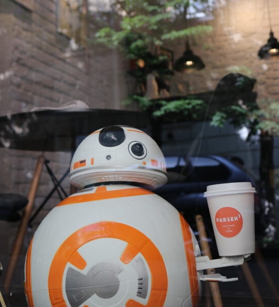 Izvan trgovine, veliki droid BB-8, mali narančasto-bijeli kružni robot iz Ratova zvijezda, drži bijelo-narančastu Parsek1 šalicu kave za ponijeti.