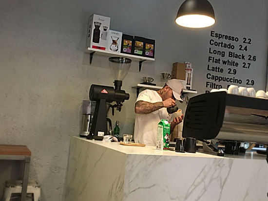 A barista in a ball cap pours a latte behind the bar at The Fix. The menu is pasted directly on the white wall behind. White shelves hold coffee bags and a chemex and other devices. The bar looks like white and gray marble. 