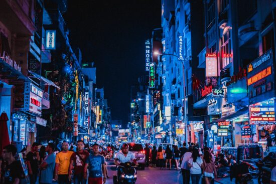 A busy city street with pedestrians, neon signs, motorcycles and cars. There are stalls with merchandise on the sidewalks and tall buildings going up, hotels and shops.