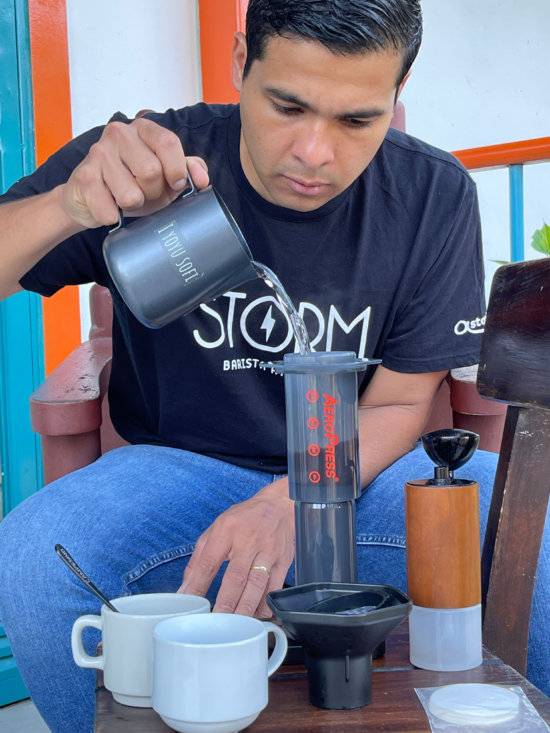 Deigo pours water from a metal pitcher into the top of an AeroPress. Two white mugs sit before him on a small table, as well as a Comandante grinder.