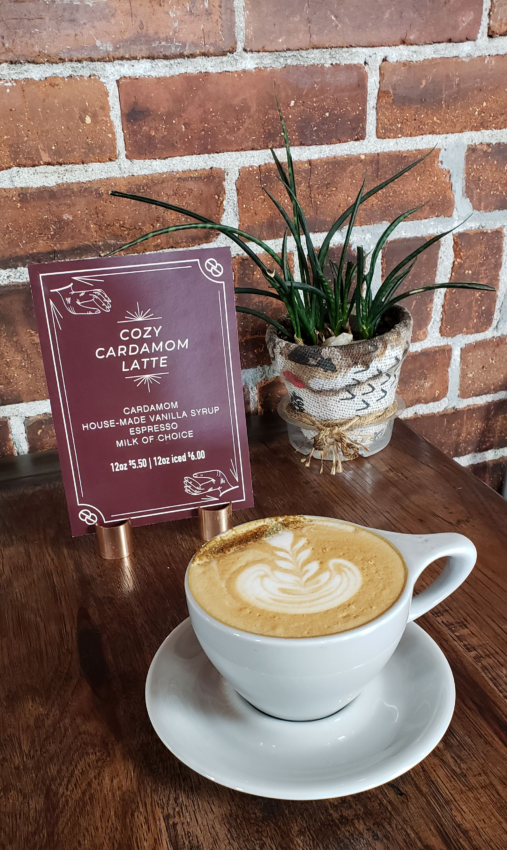 A sign for the cozy cardamom latte next to a small grass plant. In front of it is a latte mug and saucer with a rosetta and sprinkling of cardamom on top.