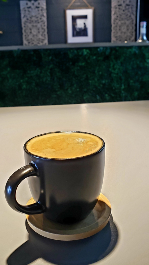 A small black mug on a wooden saucer holds a frothy cubano drink.
