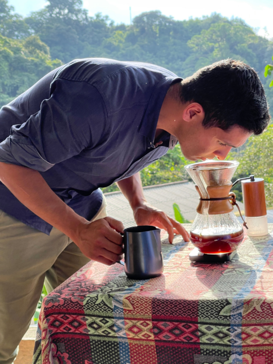 Diego snifss coffee brewing in a Chemex on a table outside. The table is covered in a woven blue and black and pink tablecloth with diamond and flower designs. He holds a metal steaming pitcher in his hand. A Comandante grinder sits behind the Chemex on the table.
