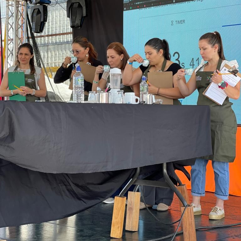 A group of four women judges sip espresso at a table with a blue tablecloth set up on the event stage. A fifth woman with a clipboard looks on. All wear green event aprons.