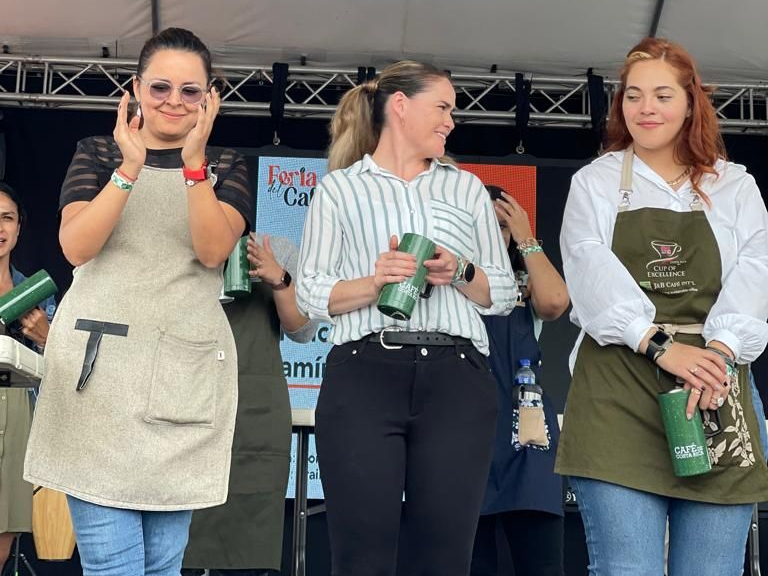 Three finalists in the Cup Tasters' champiuonship round pose for a photo. In the center is first place winner Sabrina Alvarado in a striped shirt, with blonde hair in a ponytail. The women hold green mugs. 