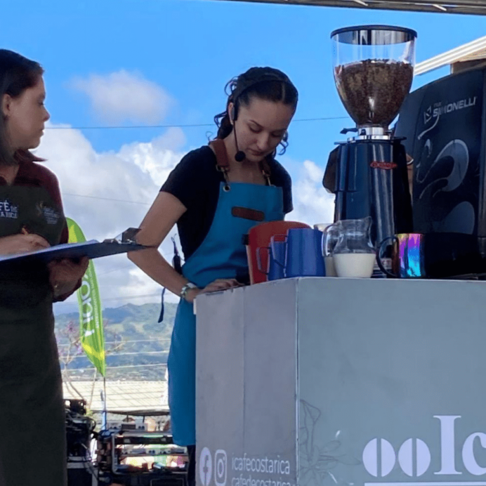 Barista Champion Sabrina Torres works at a Simonelli espresso machine on stage, while a judge watches. Sabrina wears a blue apron, has her hair tied back, and wears a microphone headset.