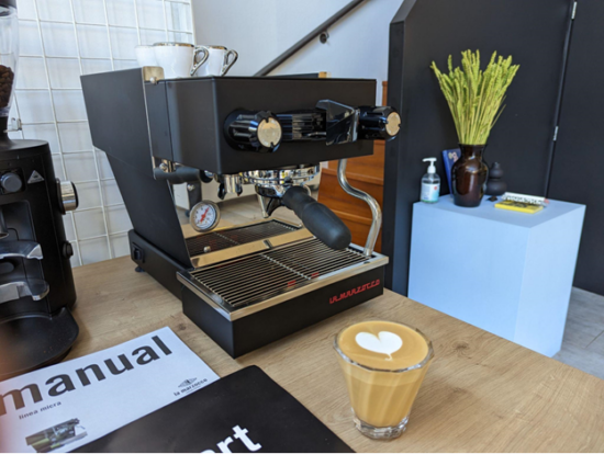 The machine on the counter with a flat white in a glass cup in front of it. There is heart latte art in the cup.
The Linea Micra is black with chrome wand and a red La Marzocco logo in all caps on the bottom right.