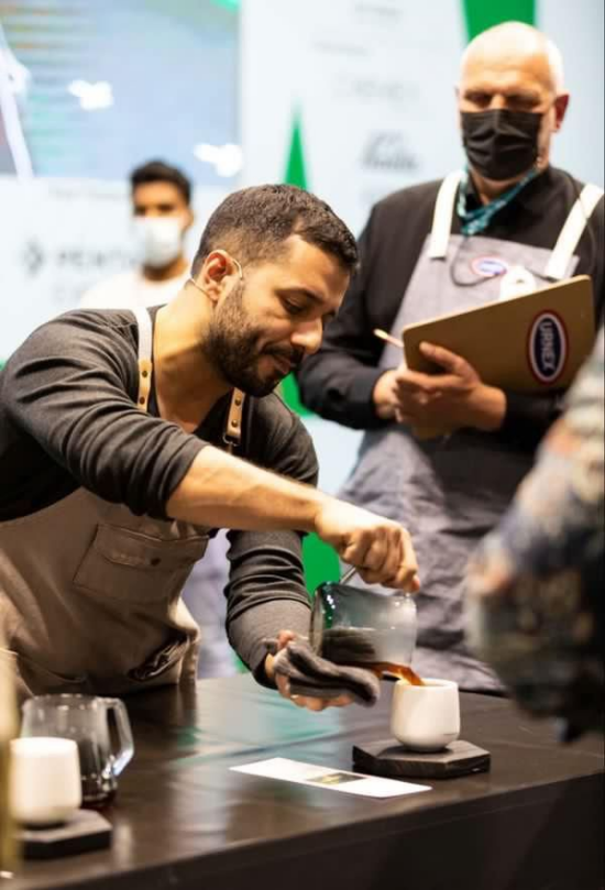 Wilford pours coffee into a cupping vessel at a competition as judges look on. He is wearing a khaki apron.