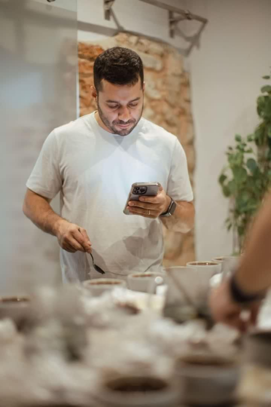 Wilford checks a recipe on his phone while cupping numerous coffees on a table. He has a close beard and white tshirt on.
