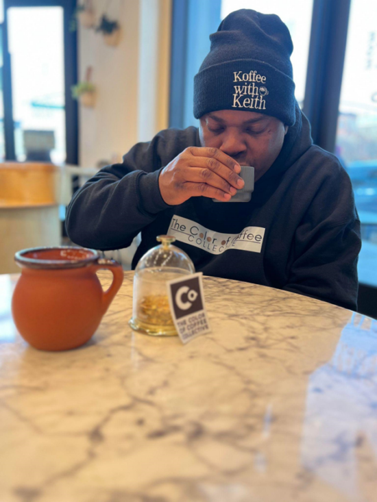 Keith sips from a gray ceramic coffee cup at a marble table. He wears a black Koffee with Keith beanie and a black Color of Coffee Collective hoodie. One the marble table is a small clay pot with a handle and a glass cloche with lights inside. A small COCC flyer leans against the globe.