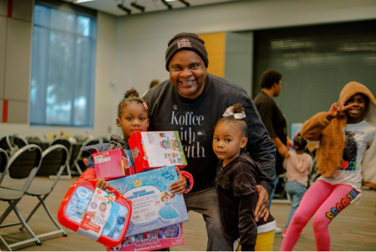 Keith is in a large room with folding chairs and large windows. He poses with two young girls holding new toys. An older girl beind them flashes a smile and a peace sign.