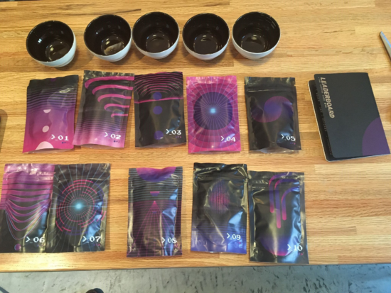 A view of the purple and black mystery coffee bags, unopened on the table, with the Leaderboard game guidebook and cupping vessels ready to be used.
