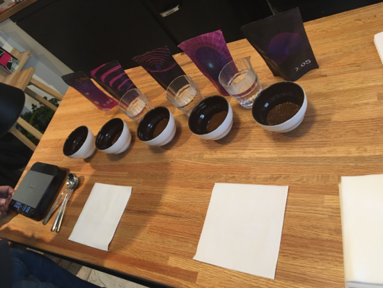 Five vessels with ground coffee await hot water. A large Acaia scale sits on the left. There are three cups of water for spoon rinsing.