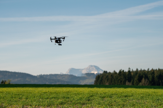 A drone hovers over grass, with a mountain, trees, and long rows of crops behind.