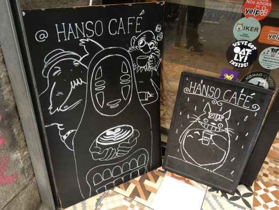 Two black sandwich boards on the street outside HanSo. One features a drawing of No Face from the anime Spirited Away, who has a mask for a face and wears a black robe. He holds a latte with rosetta art. The smaller sign next to it features Totoro of the anime MY Neighbor Totoro, who is a big cat-faced and bear-bodied beast with pointy ears. He holds a to-go cup and flashes his signature toothy grin.