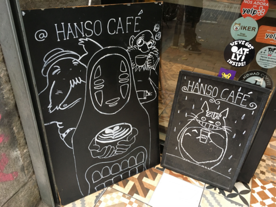 Two black sandwich boards on the street outside HanSo. One features a drawing of No Face from the anime Spirited Away, who has a mask for a face and wears a black robe. He holds a latte with rosetta art.  The smaller sign next to it features Totoro of the anime MY Neighbor Totoro, who is a big cat-faced and bear-bodied beast with pointy ears. He holds a to-go cup and flashes his signature toothy grin.