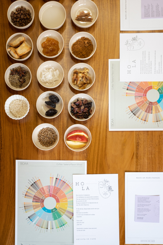An assortment of bowls containing foods for sniffing, papers with instructions, and flavor wheels. Some of the bowls contain dates, walnuts, coffee beans, star anise, and apples, to name a few.