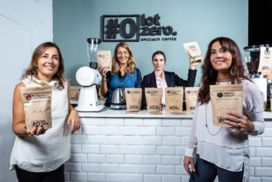 Four smiling women, the Mauro sisters, hold up coffee bags at Lot Zero. Thwy all have long hair and wear business casual attire. 