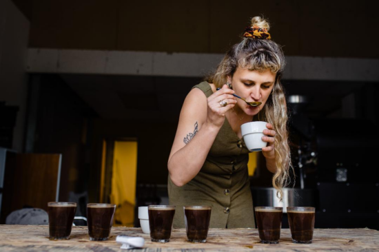 Noni sips coffee with a spoon from a cupping vessel. She has long blonde hair and wears a green dress.